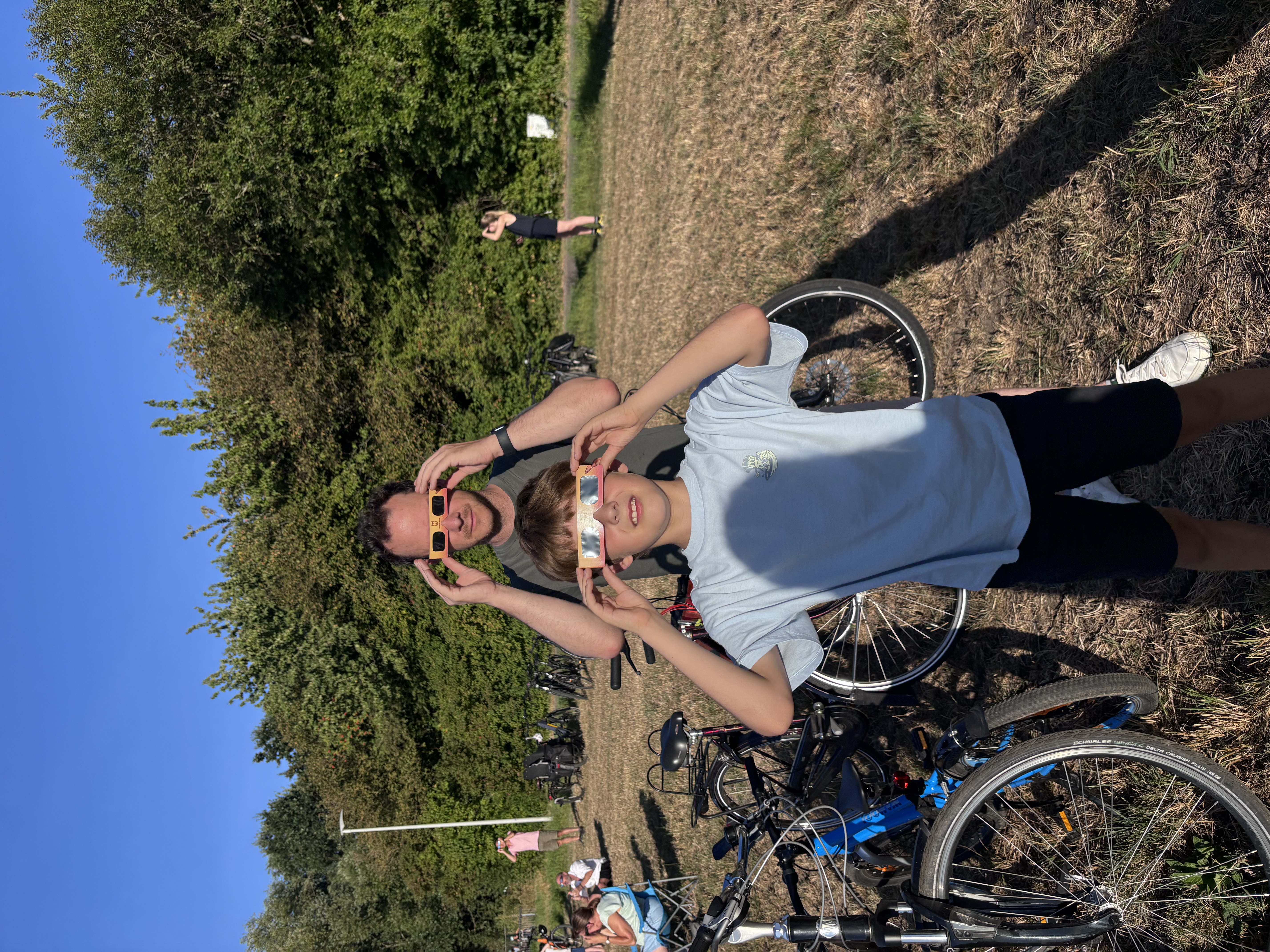 Vader en zoon met eclipsbrillen kijken naar de zon in een veld met fietsen en bomen.