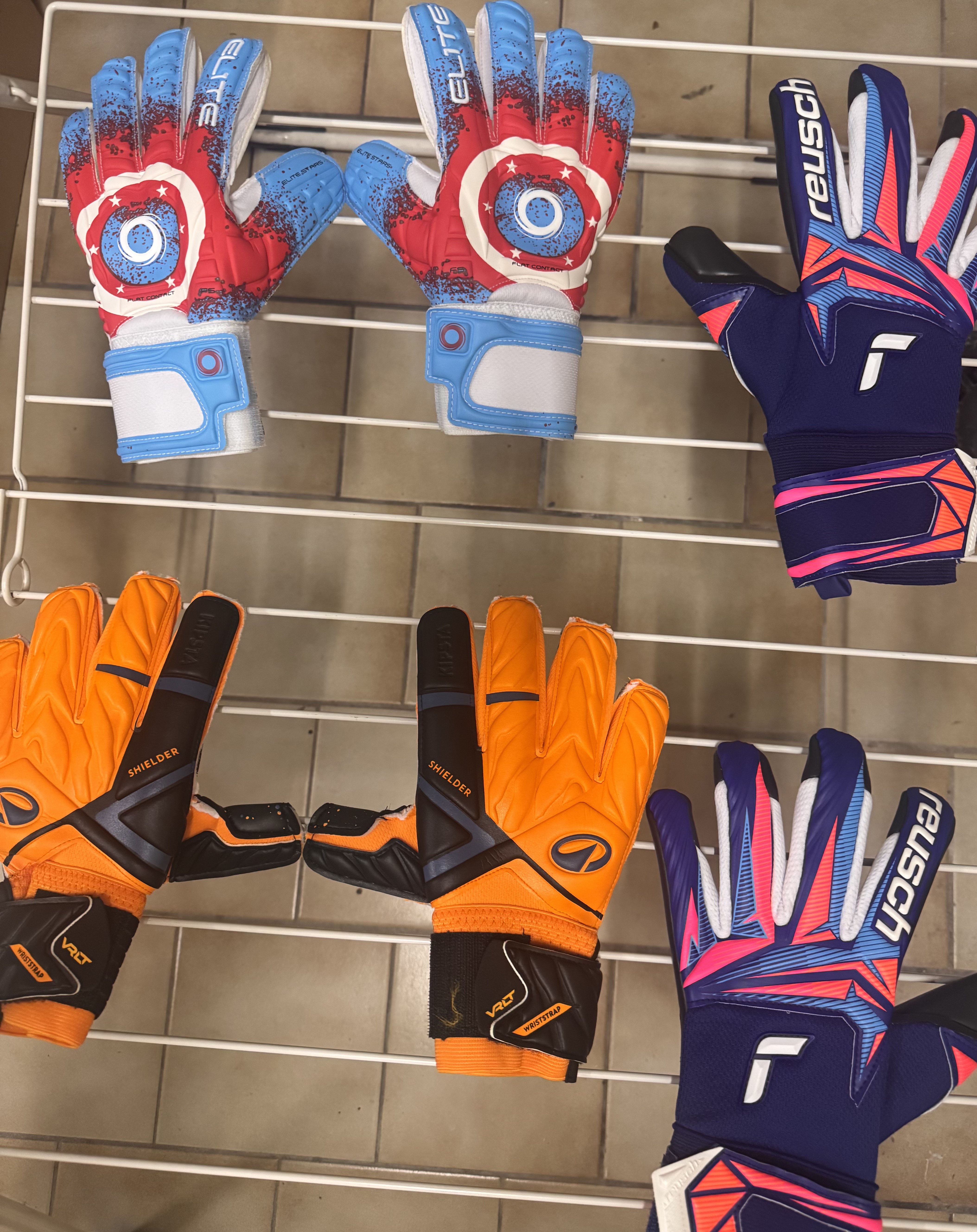 Vier kleurrijke voetbalhandschoenen in rood, blauw, oranje en paars liggen op een witte metalen droogrek.