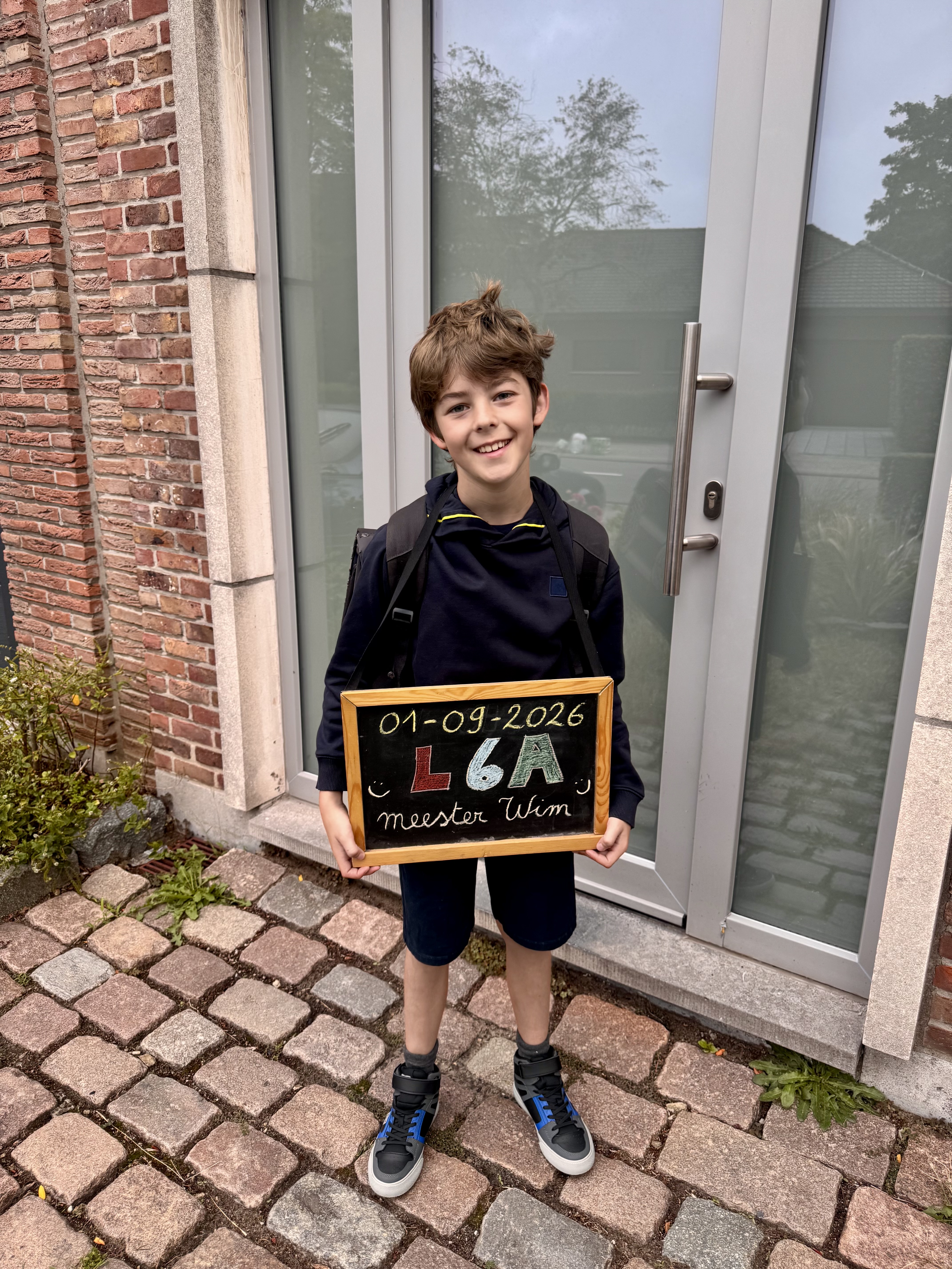 Een jongen met een rugzak en een schoolbord glimlacht voor een glazen deur, met datum en klasinformatie geschreven.