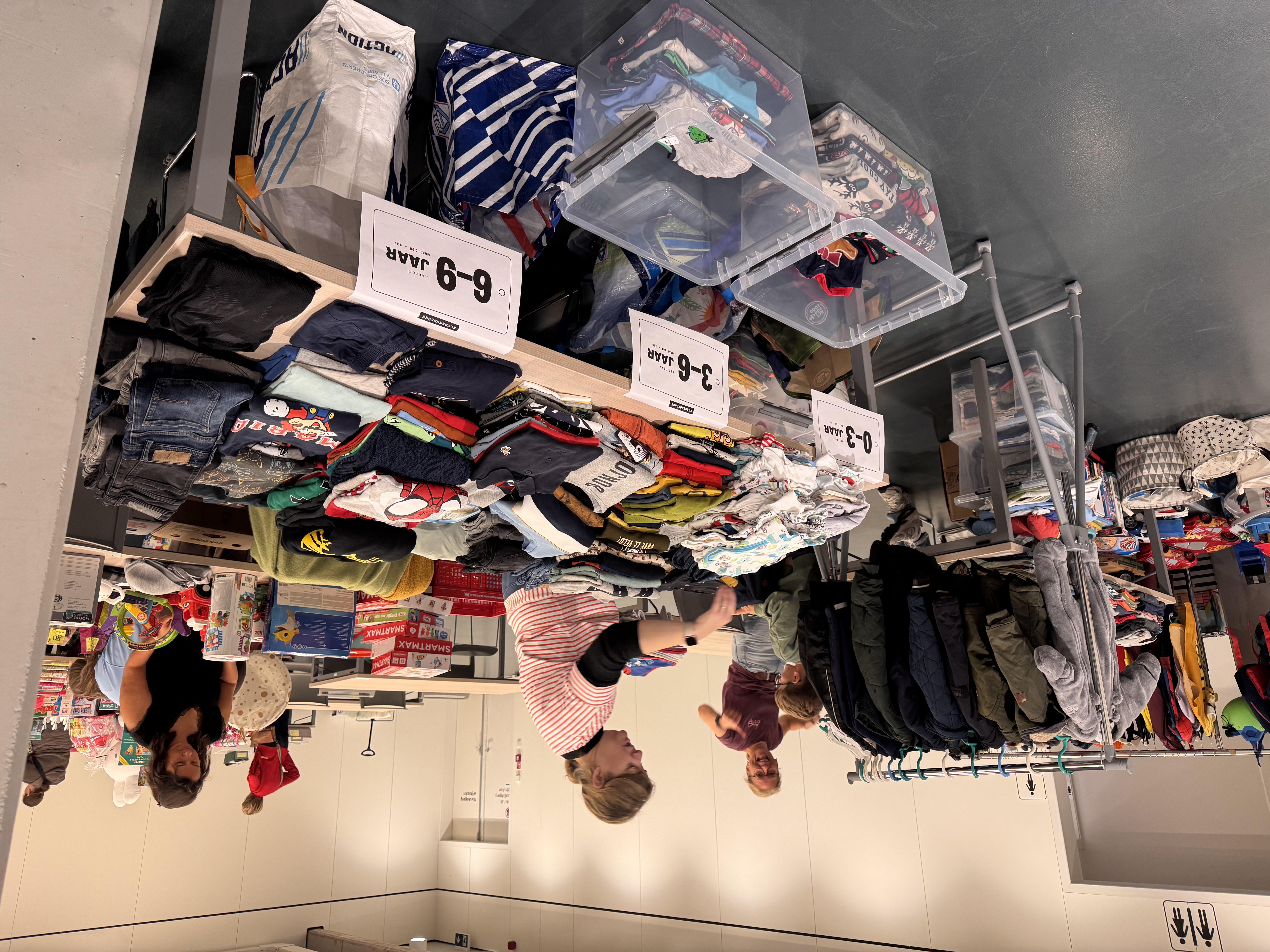 Mensen bij kledingtafels met leeftijdslabels en rekken in een zaal.